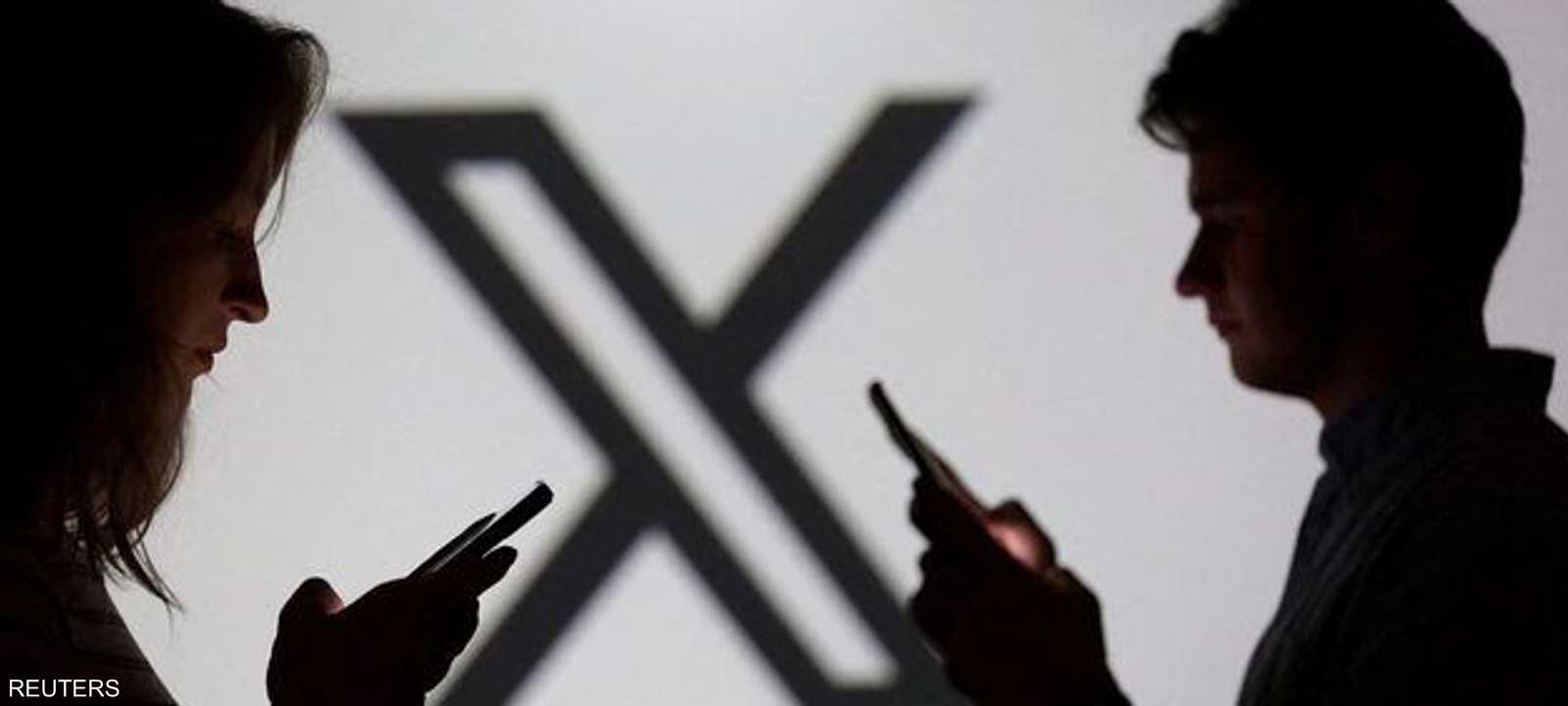 شخصان يحملان هواتف ذكية أمام شعار منصة إكس في صورة تعبيرية